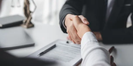 Two individuals engaging in a handshake above a document, symbolizing agreement and collaboration in a professional setting.