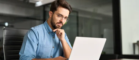 A man wearing glasses is seated at a desk, focused on his laptop while working intently.