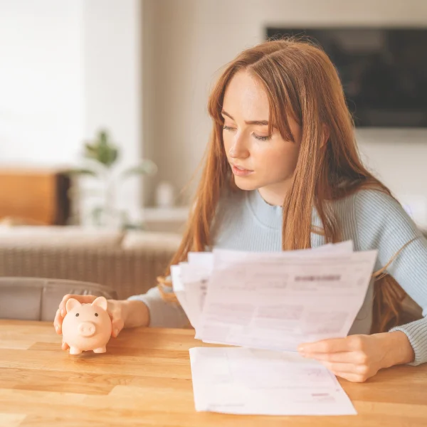 A woman examines tax documents while holding a piggy bank, reflecting on her financial situation at home.